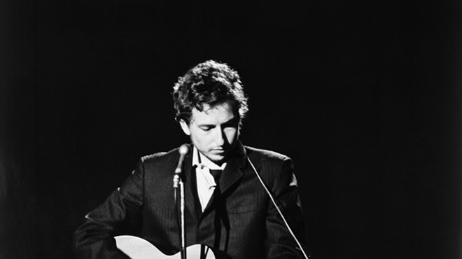 Black and white photo of Bob Dylan on stage in 1969, singing and strumming an acoustic guitar